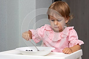 Little kid girl eating her morning porridge oatmeal with blueberries herself, sitting in baby chair in kitchen