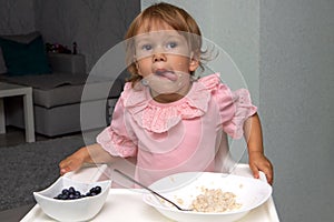 Little kid girl eating her morning porridge oatmeal with blueberries herself, sitting in baby chair in kitchen