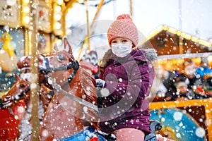 Little kid girl with medical mask on face riding on merry go round carousel horse at Christmas funfair or market. masks