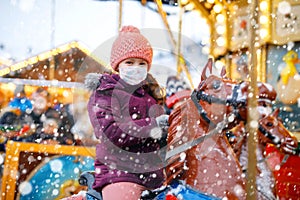 Little kid girl with medical mask on face riding on merry go round carousel horse at Christmas funfair or market. masks