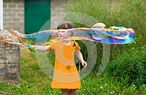 Little kid girl with pigtails in a yellow summer dress launches huge soap bubbles in the garden