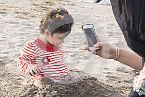 Little kid playing at beach with sand and her mother taking p