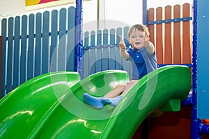 Little kid riding from childrens slides in game center and shows thumbs up. Happy little kid, children riding up, down on slide, i