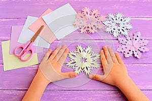 Little kid shows paper snowflakes. Children hands on lilac wooden table. Beautiful colored snowflakes diy cut from paper