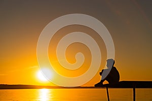 The little kid sits on a bridge and looks on the river on the background at sunset. A boy is relax near the lake