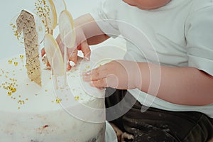 Little kid sitting near white cake, touching it with his hands and trying to taste it. The child has dirty hands and pants from th