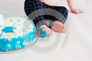 Little kid sitting near white cake, touching it with his hands and trying to taste it. The child has dirty hands and pants from th