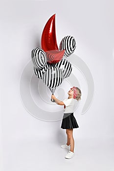 Little kid in t-shirt, red headband, black skirt and sneakers. Holding balloons, posing sideways isolated on white. Full length