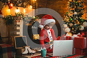 Little kid is wearing Santa clothes by his laptop. Christmas concept. Fireplace background. Christmas light.