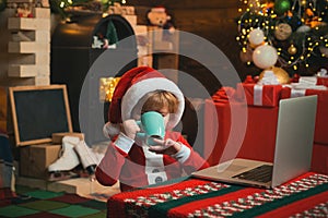 Little kid is wearing Santa clothes sitting by his laptop drinking hot Christmas drink. Fireplace background. Christmas