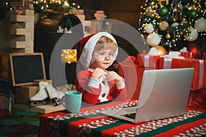 Little kid is wearing Santa clothes sitting by his laptop. Santa helper using notebook. Christmas little boy typing
