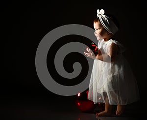 Little kid in white headband and dress, barefoot. She posing with two red balls. Twilight, black background. New Year, holidays.