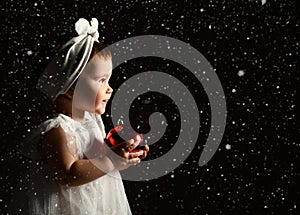 Little kid in white headband and dress, barefoot. She posing with two red balls. Twilight, black background. New Year, holidays.