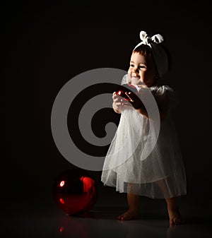 Little kid in white headband and dress, barefoot. She posing with two red balls. Twilight, black background. New Year, holidays.
