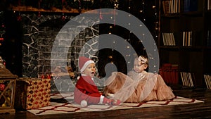 Little kids, a boy dressed as Santa Claus and a girl in a beautiful dress are playing near the New Year's tree at home.