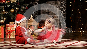 Little kids, a boy dressed as Santa Claus and a girl in a beautiful dress are playing near the New Year's tree at home.