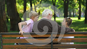 Little kids sharing secrets with grandfather resting on bench in park, back view