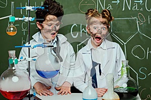 Little kids in white coats with chalkboard behind in science laboratory