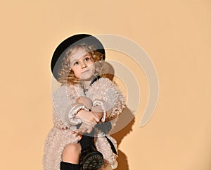 Little lady in pink faux fur coat, black hat and boots. She hugged her crossed legs while sitting against beige background