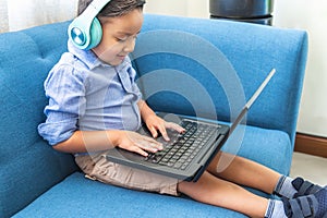 Little latin boy with headphones smiling with a laptop sitting on a blue sofa