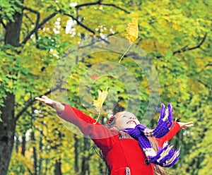 Little laughing pretty girl in red coat throws yellow leaves in autumn park