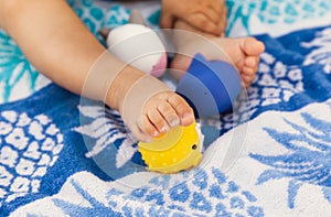 Little legs of a baby girl sitting on a beach towel and playing with rubber toys at summer time