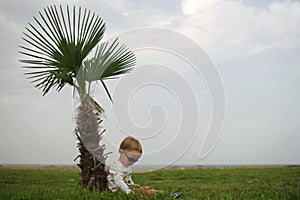 Little boy in sunglasses sitting on grass near small palm tree. View from side. Sea, horizon, cloudy sky on background