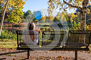 Little lonely girl with a blue balloon and a bunch of a fall leaves sits on an empty bench in a fall park.