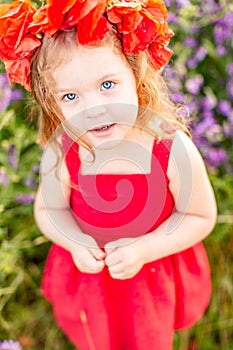 Little longhaired blonde girl in red dress at field of poppies with wreath at her head on summer sunset