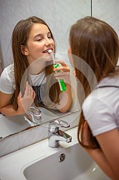 Little lovely girl brushing her teeth with electric toothbrush looking at her reflection in mirror in bathroom