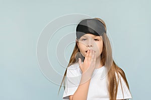 Little lovely girl wearing a sleep mask yawns over isolated blue background, studio shot, child getting ready for bed