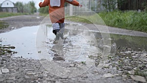 Little male child in rubber boots jogging cheerfully and falling into a puddle after rain on a rural road