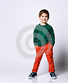 Little male in green jumper, orange pants, blue sneakers. He put hands in pockets and smiling, posing isolated on white