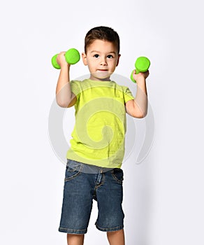 Little male in yellow t-shirt and denim shorts. He smiling, lifting green dumbbell, posing isolated on white studio background