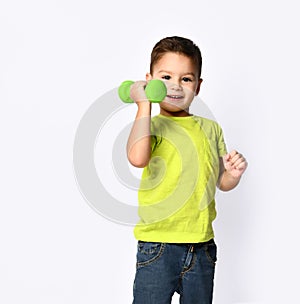 Little male in yellow t-shirt and denim shorts. He smiling, lifting green dumbbell, posing isolated on white studio background