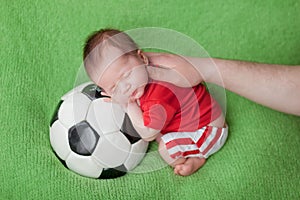 Little newborn baby sleeping on a big soccer ball in a red and white soccer player suit on a green background