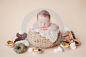 A little newborn baby sleeps in a straw basket on a beige background, there are many donuts, cakes and sweets