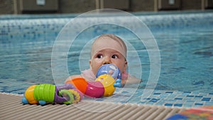 A little newborn girl is swimming in the pool and playing with a colorful toy.