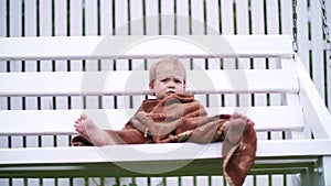 A little one-year-old girl, wrapped in a towel, with a dirty face, lonely sitting on a swing in the garden, in the