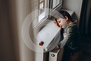 Little, pensive, two years old boy sitting by the window and playing with toy children& x27;s car