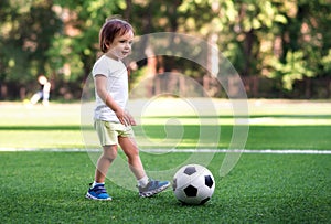 Little player: toddler boy in sports uniform playing footbal at soccer field in summer day outdoors. Child ready to kick ball