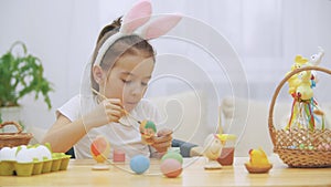 Little playful girl, wearing bunny ears on her head chose an orange colour to paint an egg. Girl is chewing and outting
