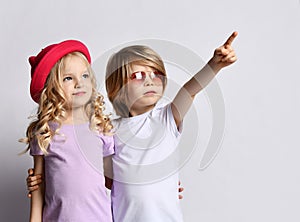 Little positive blond baby girl and boy in stylish clothing standing, hugging each other and pointing at something above