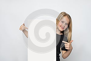 Little positive girl shows index finger at blank poster against white background