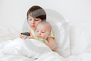 Little preschool boy laying in bed with his baby brother, playing on phone