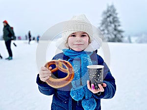 Little preschool girl drinking hot chocolate and eating pretzel in winter forest. Happy healthy child with cup of