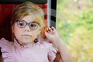 Little preschool girl with glasses sitting in train and looking out of window while moving. Happy child , smiling child