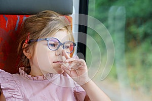 Little preschool girl with glasses sitting in train and looking out of window while moving. Happy child , smiling child