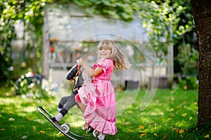 Little preschool girl hugging with rocking horse toy. Happy child in princess dress on sunny summer day in garden. Girl