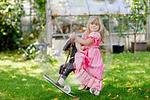 Little preschool girl hugging with rocking horse toy. Happy child in princess dress on sunny summer day in garden. Girl